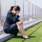A woman in a business suit sits on the grass by a fence, head in her hand, appearing stressed or upset—with the urban background hinting at workplace sexual harassment: signs and the need for evidence & legal steps.
