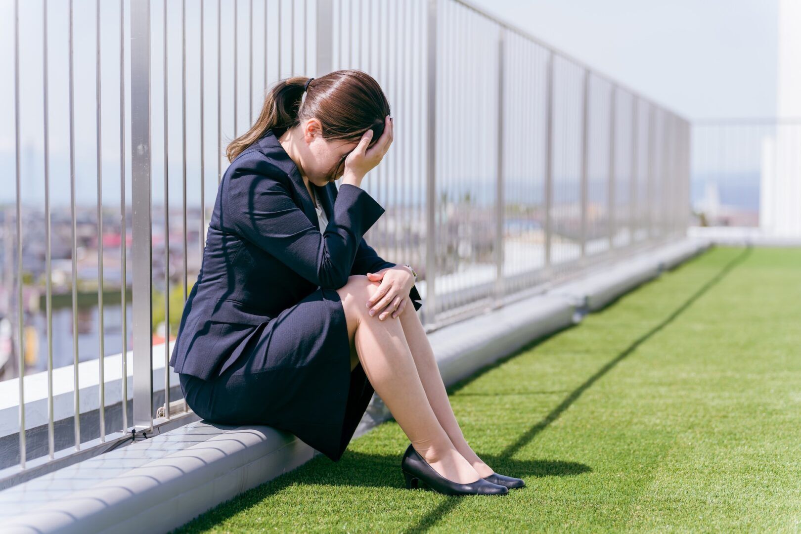 A woman in a business suit sits on the grass by a fence, head in her hand, appearing stressed or upset—with the urban background hinting at workplace sexual harassment: signs and the need for evidence & legal steps.