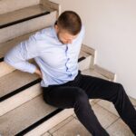A man in a light blue shirt and black pants sits on indoor stairs, holding his lower back in pain—raising the question: Can You Sue for a Slip and Fall? WV Legal Guide may provide the answers you need.