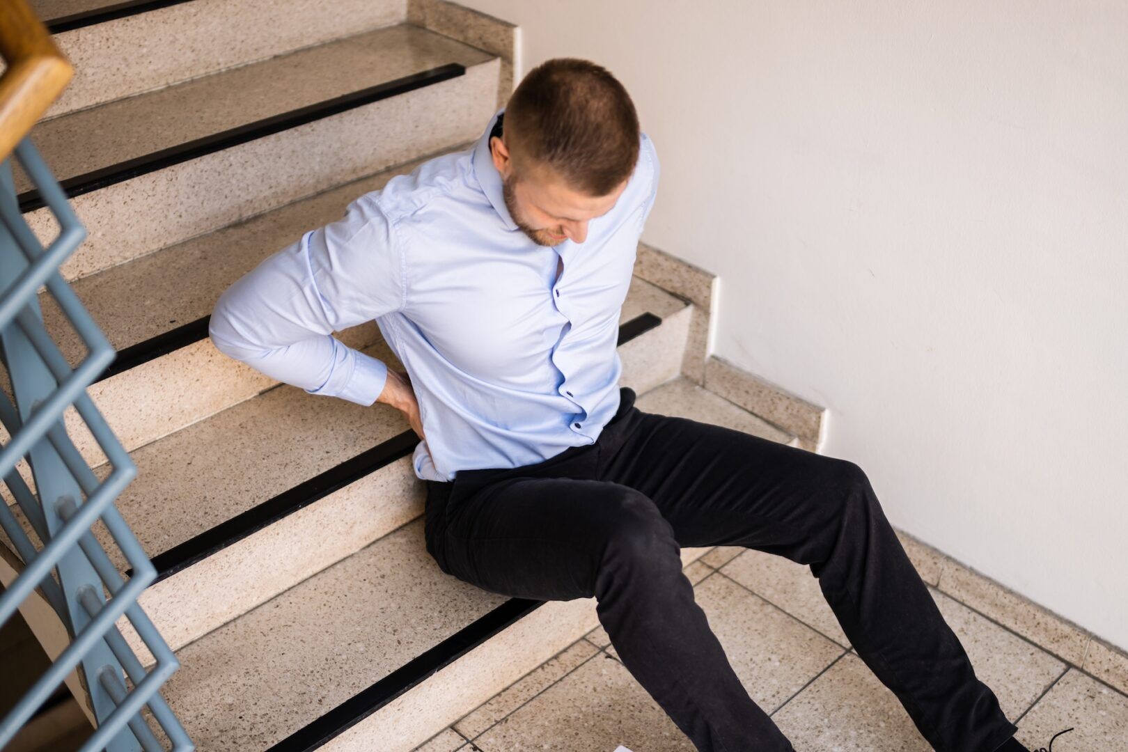 A man in a light blue shirt and black pants sits on indoor stairs, holding his lower back in pain—raising the question: Can You Sue for a Slip and Fall? WV Legal Guide may provide the answers you need.