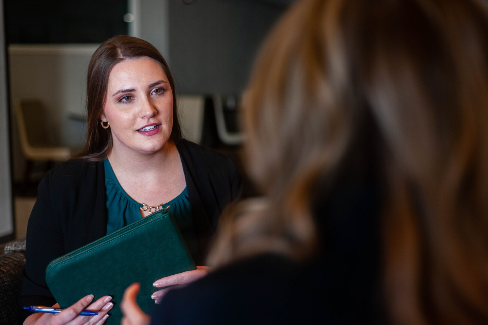 A woman with long brown hair, holding a green folder and pen, listens attentively to another person whose back is to the camera in a professional indoor setting.