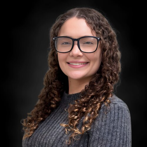 A woman with long, wavy dark hair wearing a light-colored, short-sleeved top smiles in front of a dark, blurred background.