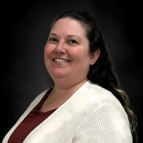 A woman with long, wavy dark hair wearing a light-colored, short-sleeved top smiles in front of a dark, blurred background.