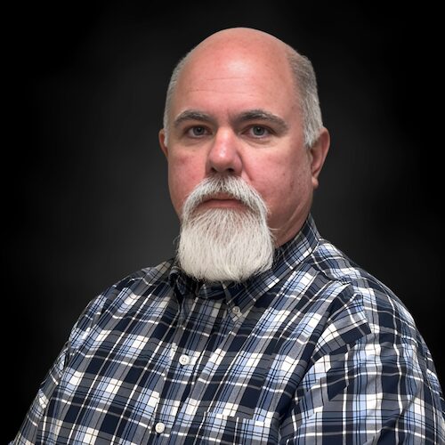 A woman with long brown hair and glasses smiles at the camera. She is wearing red floral earrings and a pink patterned top, set against a plain black background.