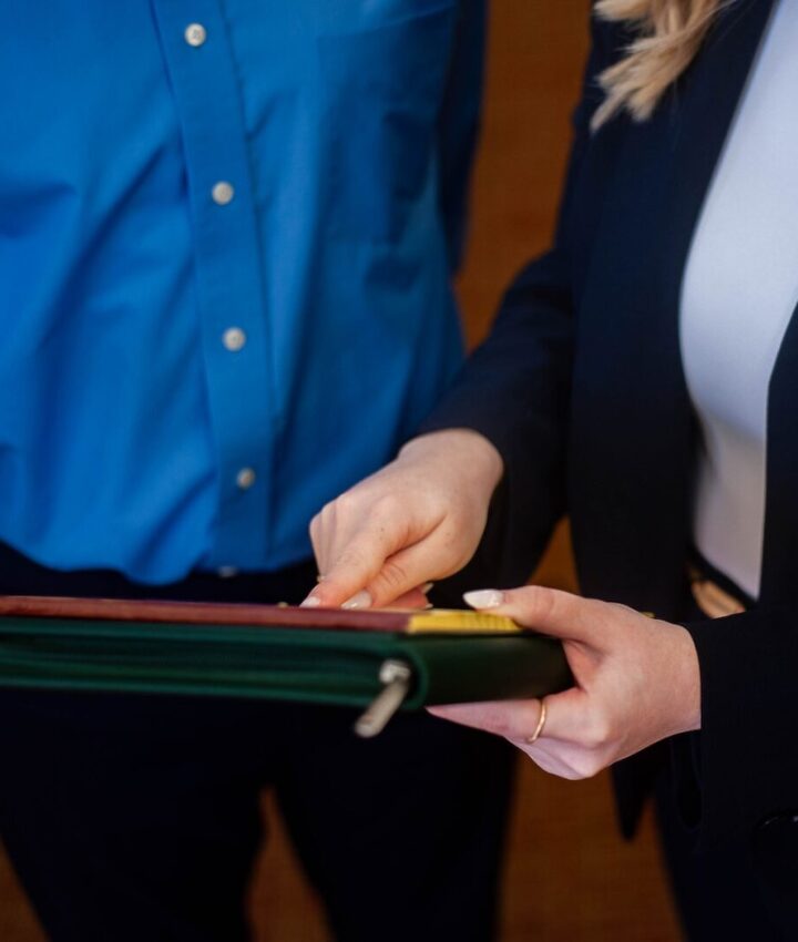 Two people stand close together, one in a blue shirt and the other in a black blazer. The person in the blazer points to a document or binder held between them. Their faces are not visible.