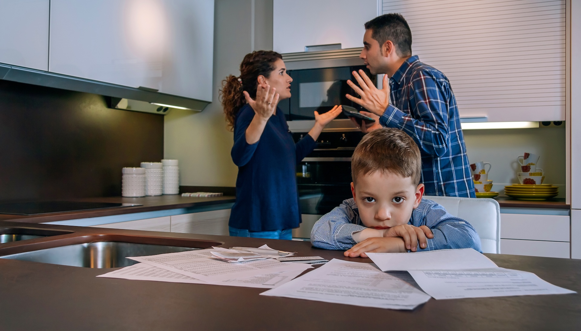A young boy looks sad at a kitchen table covered with papers while a man and woman argue in the background, both gesturing with frustration.