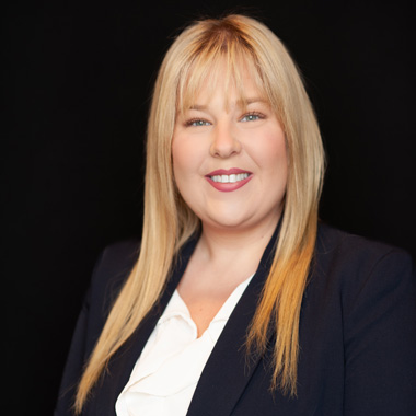 Woman with straight blonde hair wearing a navy blazer over a white blouse, smiling and posing in front of a solid black background.