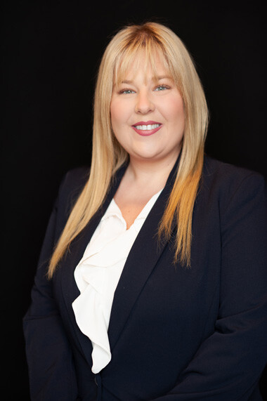 A woman with straight blonde hair wearing a navy blazer and white blouse smiles at the camera against a plain black background.