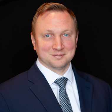A man with short light brown hair wearing a dark blue suit, white shirt, and patterned tie, smiling in front of a plain black background.