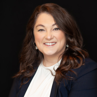 A woman with long brown hair, wearing a navy blazer and white blouse, smiles at the camera against a plain black background.
