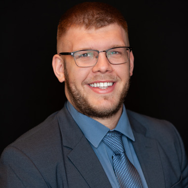 A man with short hair, a beard, and glasses smiles at the camera. He is wearing a blue dress shirt, a patterned tie, and a dark suit jacket against a plain black background.