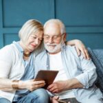 An elderly couple sits closely together on a sofa, smiling while looking at a tablet. The woman has her arm around the man, and they appear happy and relaxed in their cozy, blue-themed living room as they research grandparents rights West Virginia.