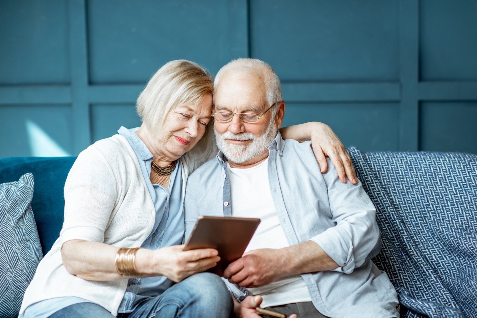 An elderly couple sits closely together on a sofa, smiling while looking at a tablet. The woman has her arm around the man, and they appear happy and relaxed in their cozy, blue-themed living room as they research grandparents rights West Virginia.