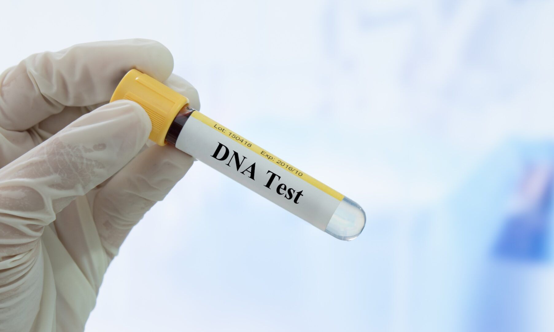 A gloved hand holds a test tube labeled "DNA Test" containing a blood sample, used to establish paternity in West Virginia, with a blurred clinical background.