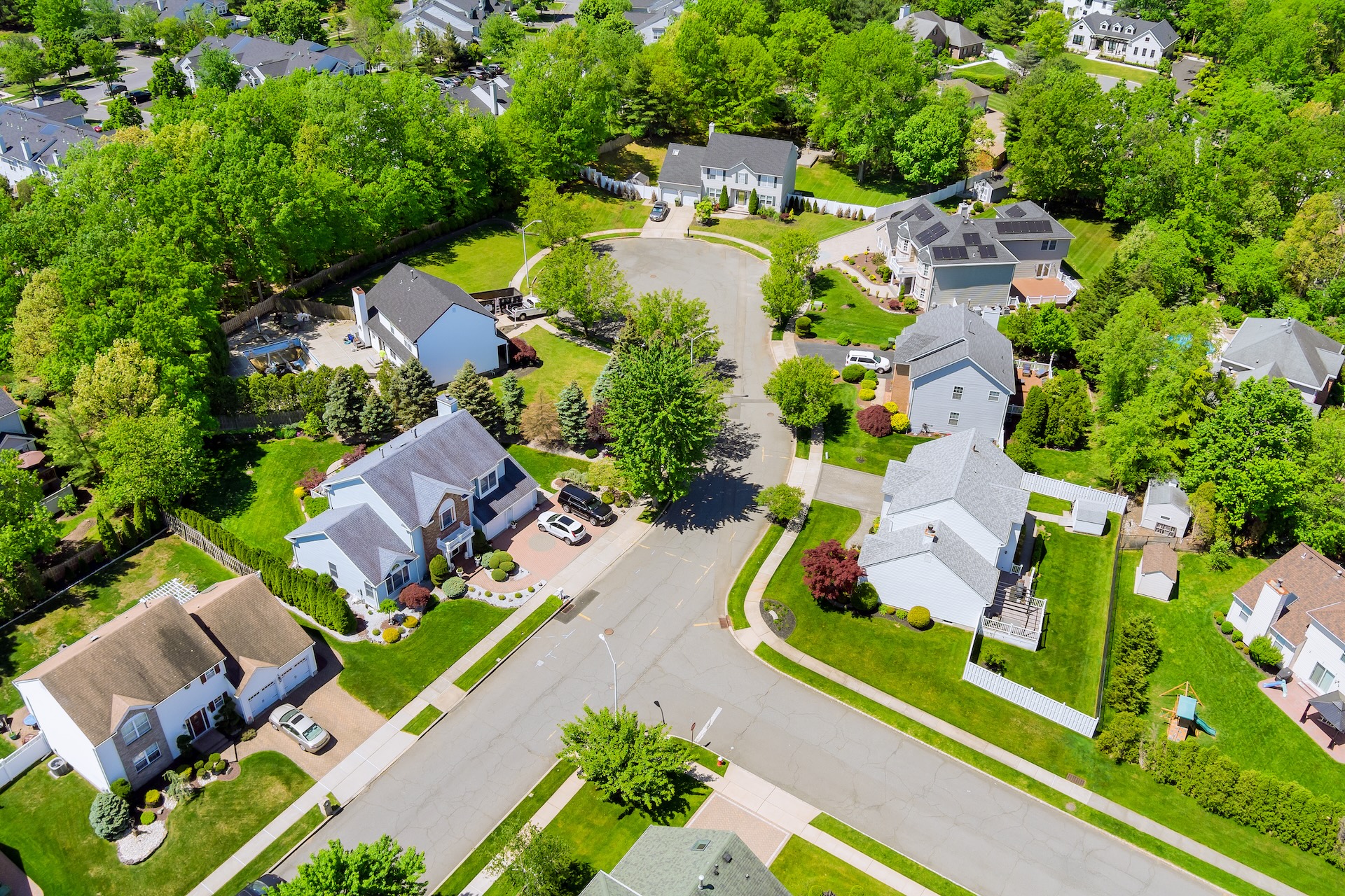 Aerial view of a suburban neighborhood with tree-lined streets, single-family homes, neatly manicured lawns, and cars parked in driveways on a sunny day.