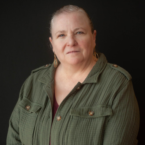 A middle-aged woman with light skin and gray hair pulled back, wearing gold hoop earrings and an olive green button-up jacket, stands in front of a plain black background.
