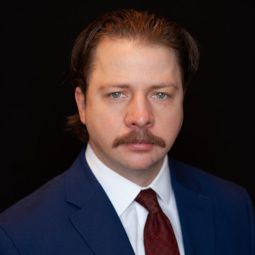 A man with light skin, light brown hair, and a mustache wearing a blue suit, white shirt, and red patterned tie, poses against a black background.