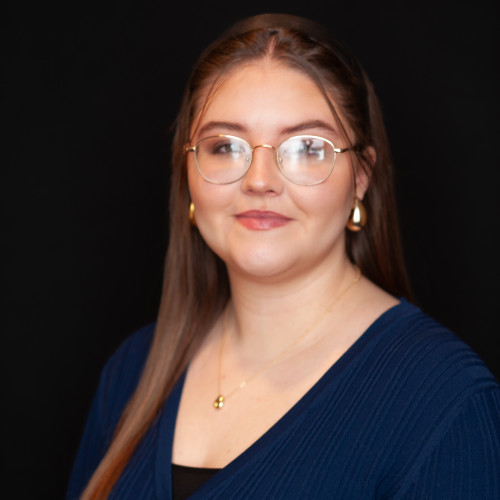 A young woman with long brown hair and glasses, wearing a navy blue top, gold hoop earrings, and a gold necklace, poses in front of a plain black background.