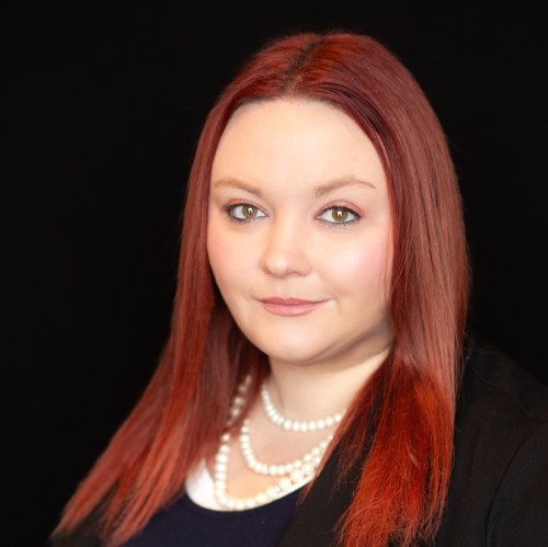 Woman with straight, long red hair wearing a black blazer, navy top, and layered pearl necklace, poses against a solid black background with a neutral expression.
