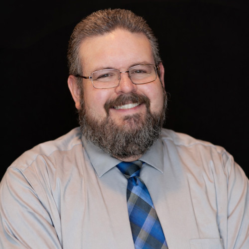 A man with glasses, a thick beard, and short hair smiles at the camera. He is wearing a light gray shirt and a blue plaid tie, set against a plain black background.