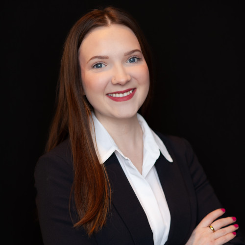 A woman with long brown hair, wearing a black blazer and white shirt, stands in front of a black background, smiling confidently with her arms crossed.