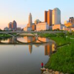 A city skyline with tall buildings reflects in a calm river at sunset. A bridge spans the water, while a person in a red shirt stands on rocks by the grassy riverbank, quietly fishing and reflecting much like one awaiting a Stark County Family Court custody decision.