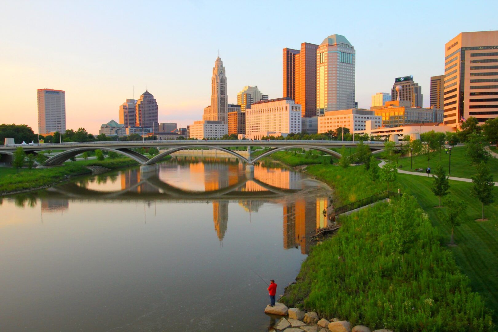 A city skyline with tall buildings reflects in a calm river at sunset. A bridge spans the water, while a person in a red shirt stands on rocks by the grassy riverbank, quietly fishing and reflecting much like one awaiting a Stark County Family Court custody decision.