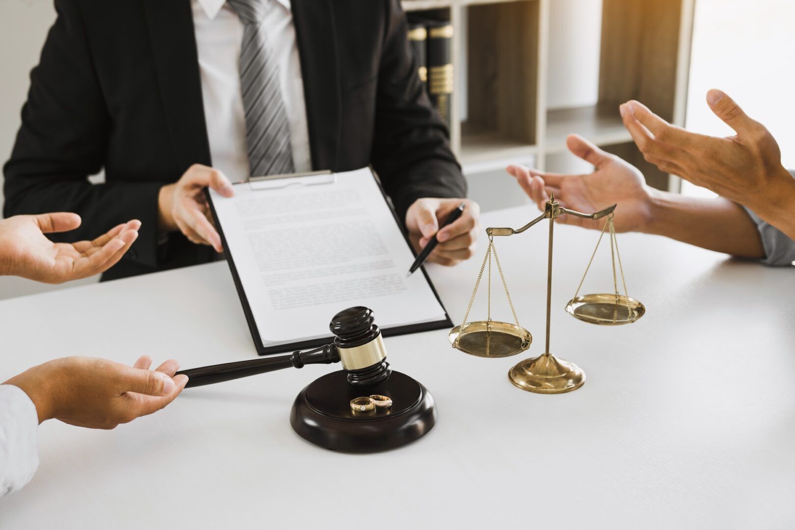 Three people are seated at a desk with legal documents, a gavel, two wedding rings, and balancing scales, suggesting a legal discussion about divorce vs dissolution in Ohio or a potential settlement.