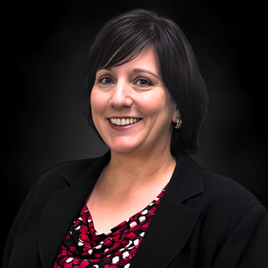 Smiling woman with short dark hair wearing a black blazer and a red patterned top, posing in front of a plain black background.