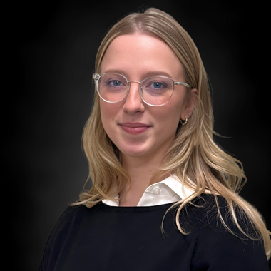 A young woman with long blonde hair, wearing clear glasses, a white collared shirt, and a black sweater, poses in front of a dark, blurred background.
