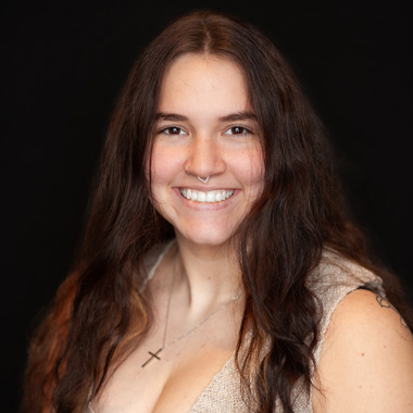 A woman with long brown hair smiles at the camera. She is wearing a beige top, a cross necklace, and has a nose ring. The background is plain black.