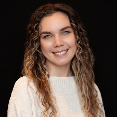 A woman with long, wavy light brown hair and blue eyes smiles at the camera. She is wearing a cream-colored sweater and is posed against a plain black background.