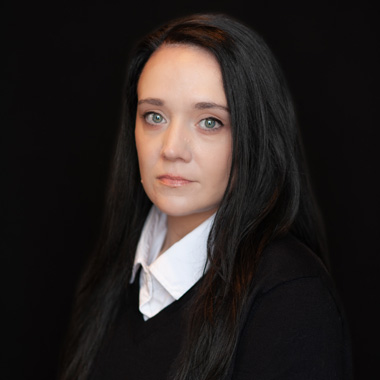 A woman with long straight black hair, light eyes, and fair skin wears a white collared shirt under a black sweater. She poses against a plain black background, looking slightly to the side with a neutral expression.