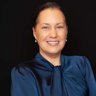 A woman with dark hair pulled back, wearing pearl earrings and a shiny blue blouse with a bow, smiles in front of a plain black background.