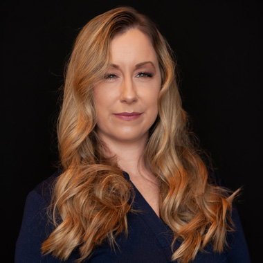 A woman with long, wavy blonde hair wearing a dark blue top stands against a plain black background, looking confidently at the camera with a slight smile.