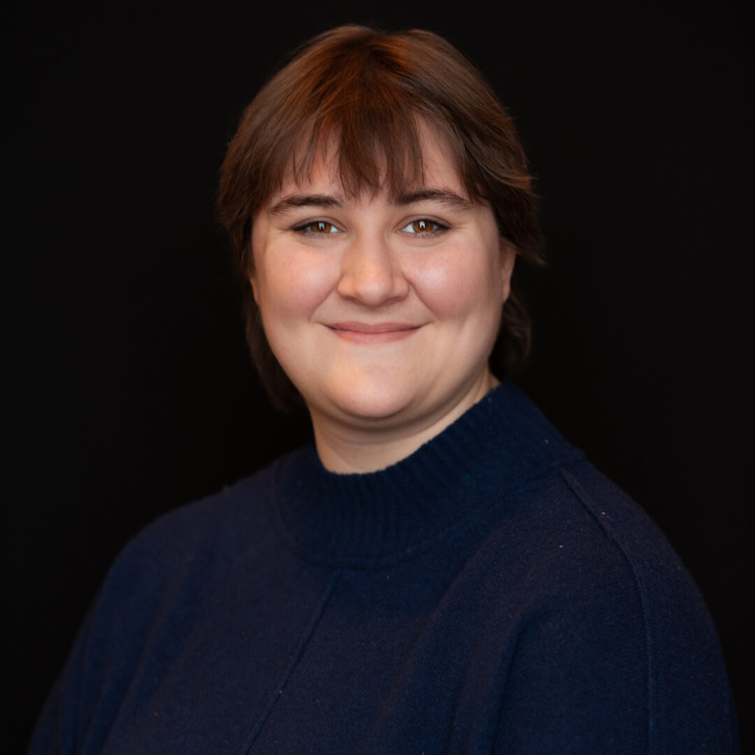A person with short brown hair and bangs smiles gently at the camera. They are wearing a dark blue sweater and are posed against a plain black background.