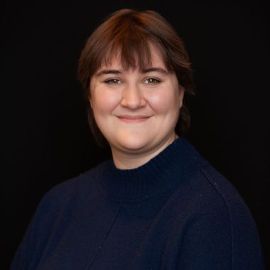 A person with short brown hair and a navy blue sweater smiles softly against a plain black background.