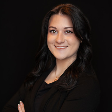 A young woman with long dark hair, wearing a black blazer and top, smiles with arms crossed against a solid black background.