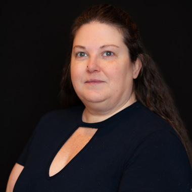 A woman with long brown hair, wearing a black top with a keyhole neckline, poses against a plain black background, looking directly at the camera with a neutral expression.