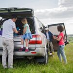 A family is parked in a grassy field with an open SUV, perhaps discussing relocate out of state child custody WV. A man helps a young girl on the back while a woman and boy stand by the side door, beneath the partly cloudy sky and scenic green backdrop.