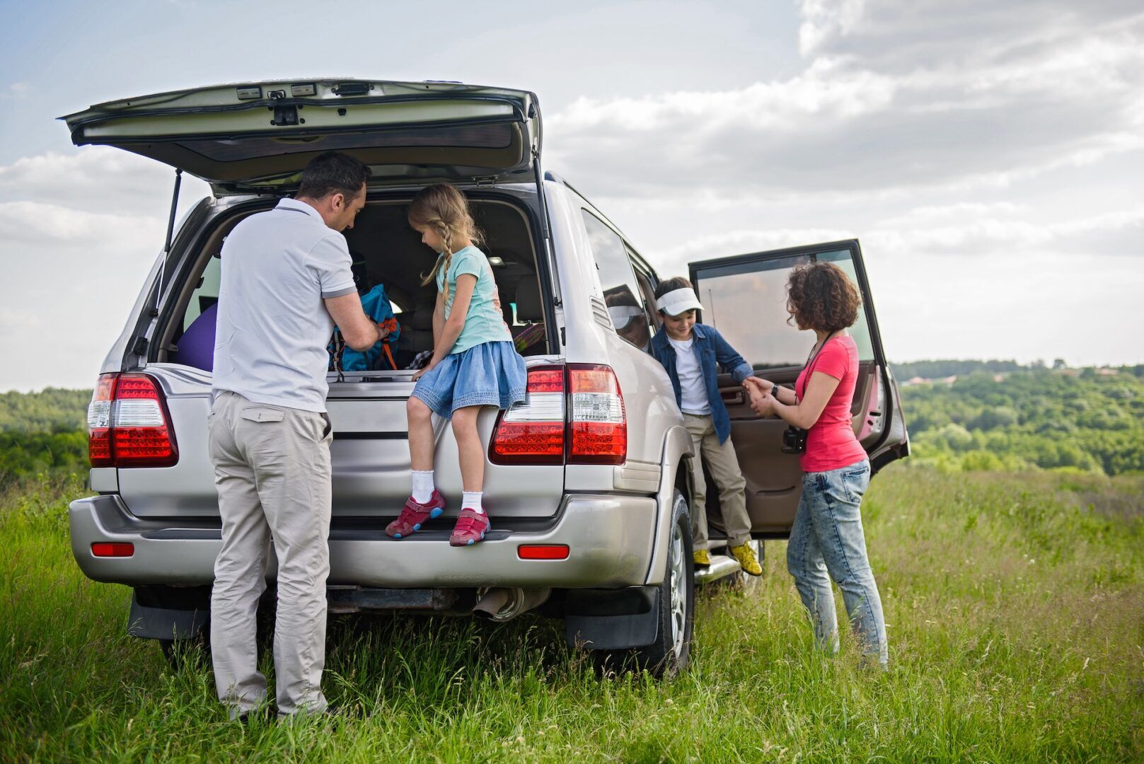 A family is parked in a grassy field with an open SUV, perhaps discussing relocate out of state child custody WV. A man helps a young girl on the back while a woman and boy stand by the side door, beneath the partly cloudy sky and scenic green backdrop.