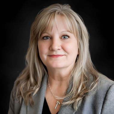 A woman with long, light blonde hair and blue eyes, wearing a gray blazer and a necklace with two pendants, smiles softly against a dark background at Travis Britton Klie Law Offices.
