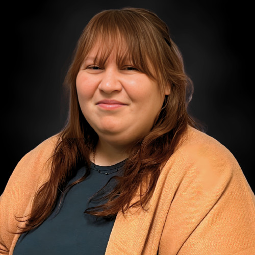 A woman with long brown hair and bangs smiles slightly. She is wearing a dark top and a tan shawl, posing in front of a dark, blurred background.