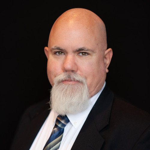 A middle-aged man with a bald head and full white beard wears a suit, white shirt, and striped tie, posing in front of a black background with a neutral expression.