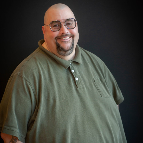 A smiling man with a shaved head, glasses, and goatee wears a green polo shirt and stands in front of a dark background.
