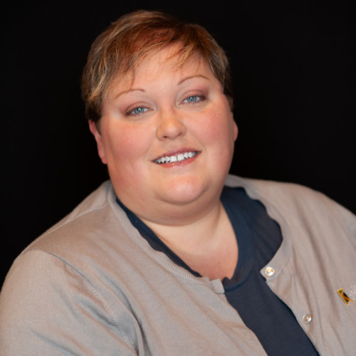 A person with short light brown hair smiles at the camera. They are wearing a beige cardigan over a dark top, with a black background behind them.