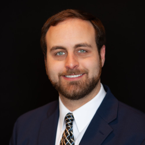 A man with short brown hair, a beard, and blue eyes is smiling. He is wearing a dark blue suit, white shirt, and patterned tie, posed in front of a plain black background.