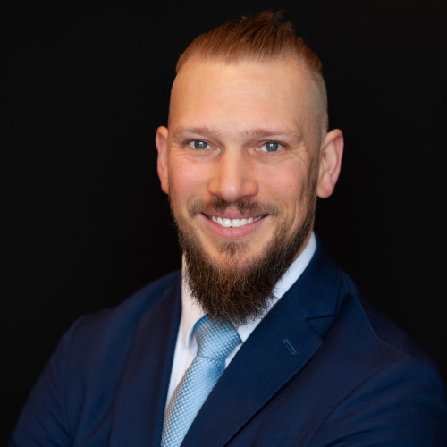 A smiling man with a long beard and blonde hair pulled back, wearing a blue suit, light blue shirt, and light blue tie, poses against a plain black background.