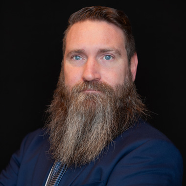 A man with a long, full brown and gray beard and slicked-back hair, wearing a dark suit jacket and a patterned tie, poses against a black background.