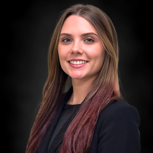 A young woman with long, straight brown hair and an ombre of darker ends, wearing a black blazer and smiling, posed against a plain dark background.